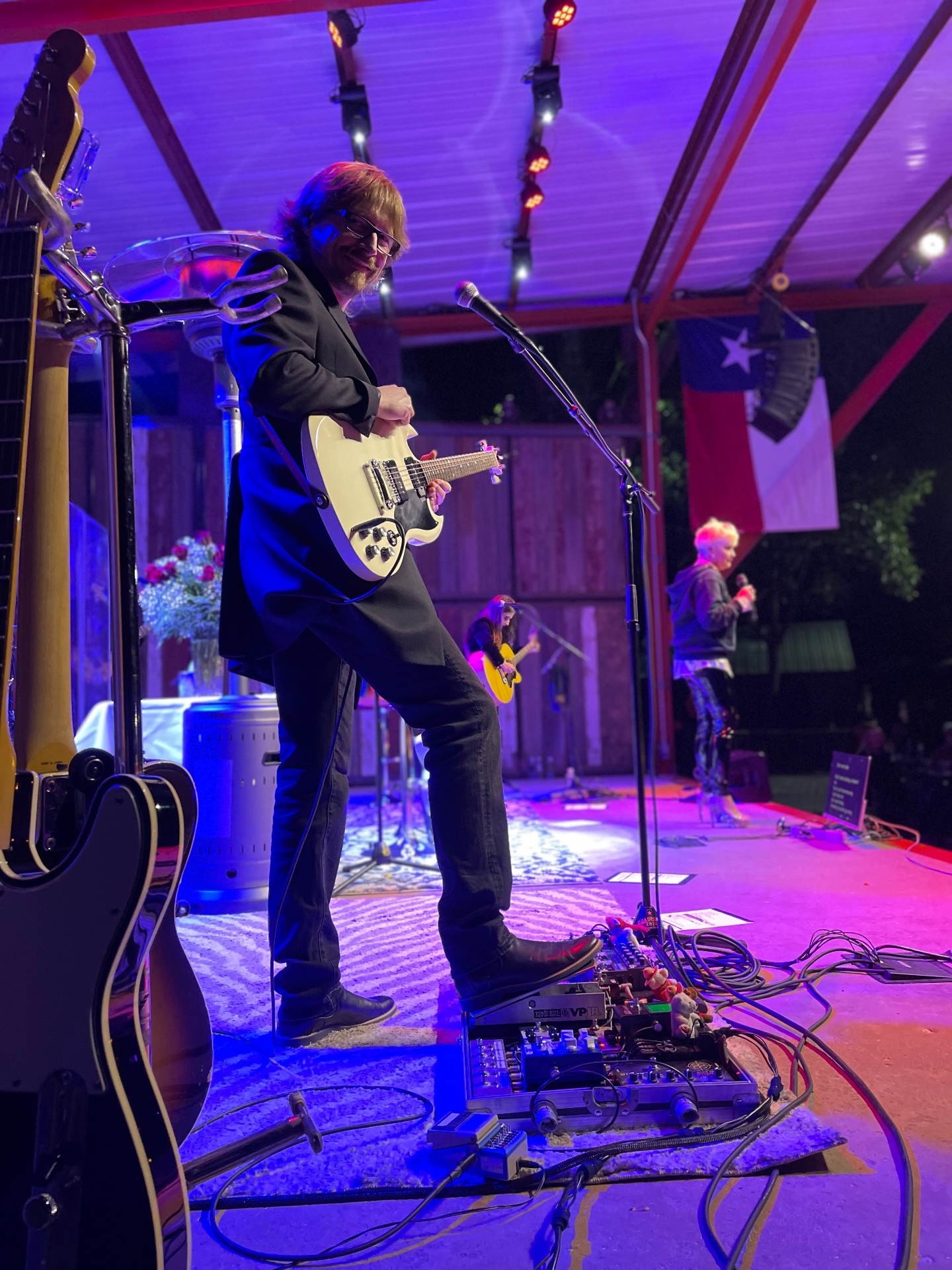 Guitarist on stage with pedalboard, vocal mic, and moving-head lighting overhead